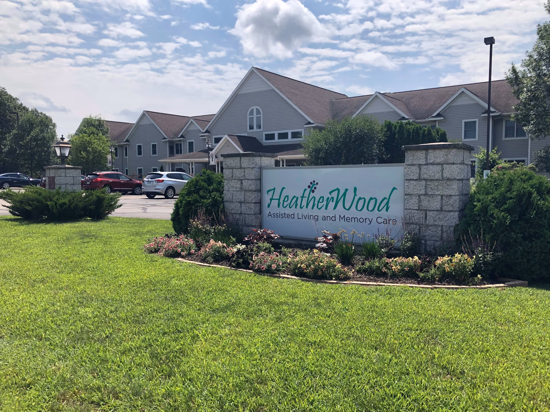 Exterior view of HeatherWood Assisted Living and Memory Care facility with a large sign in front surrounded by flowers and greenery. The building is a two-story structure with multiple windows and a sloped roof. Several cars are parked in the parking lot nearby under a partly cloudy sky.