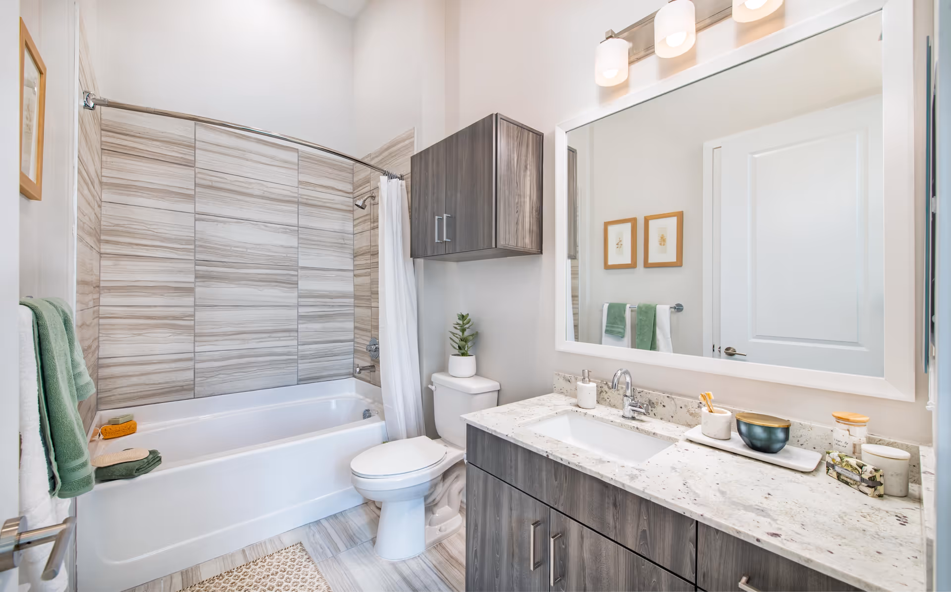 Bright modern bathroom with a tub/shower and tiled surround, toilet, and a vanity with wood cabinets and a large mirror.