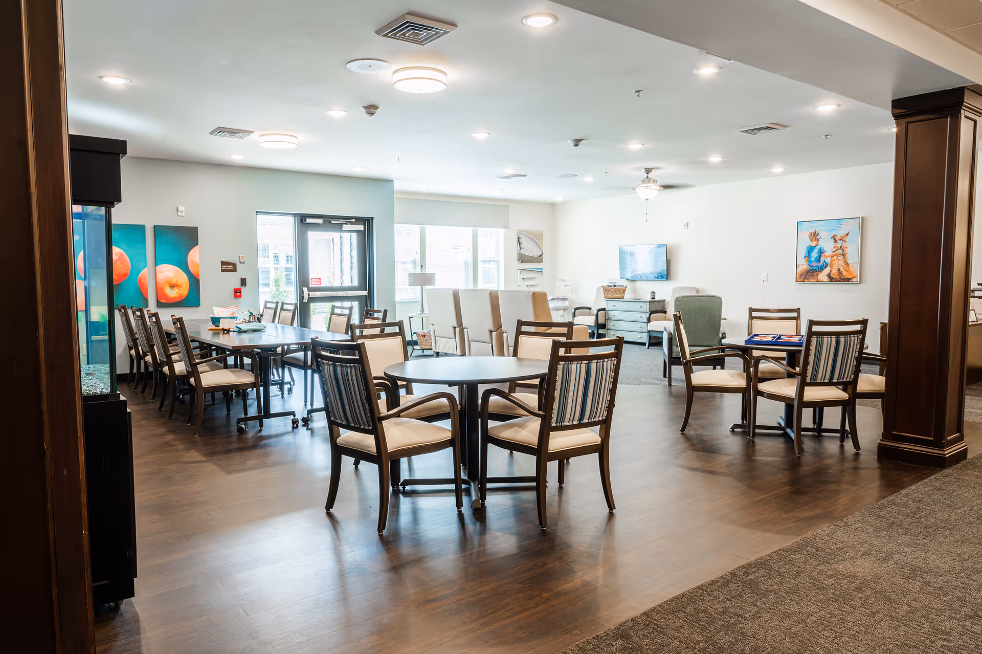 A spacious and well-lit common area with multiple tables and chairs arranged for dining or social activities. The room features wooden flooring, large windows allowing natural light, colorful artwork on the walls, and a television mounted on the far wall. There are also comfortable armchairs and a cabinet beneath the TV.