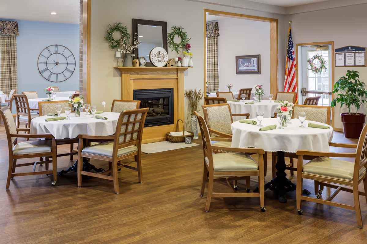 A dining room in Glenwood Place featuring round tables covered with white tablecloths, each set with green napkins, glassware, and floral centerpieces. Wooden chairs with cushioned seats surround the tables. The room has a wooden floor, a fireplace decorated with wreaths and a sign that reads 'Gather around the table,' and an American flag near a glass door. A large wall clock and framed artwork are visible in the background.