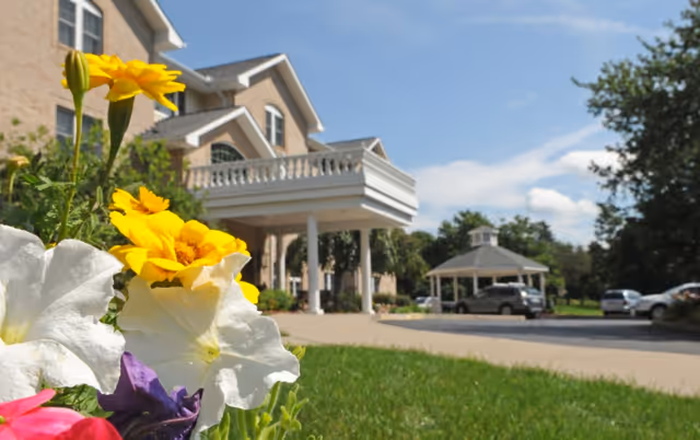 Close-up of colorful flowers in the foreground with a senior living facility building and a covered driveway entrance in the background under a blue sky with some clouds.