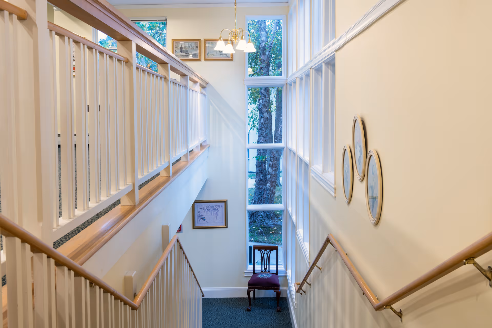 Interior view of a stairwell with wooden handrails and white balusters. There is a large window at the landing letting in natural light and showing trees outside. The walls are painted light yellow and decorated with framed pictures and three oval wall hangings. A wooden chair with a cushioned seat is placed at the bottom of the stairs.