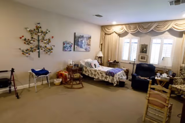 A cozy bedroom in a senior living facility with a single bed covered in a patterned bedspread, a wooden rocking chair, a recliner, and another armchair. The room has beige walls decorated with a metal tree wall art and framed pictures. There are three windows with white shutters and beige curtains, and various small tables with lamps and personal items. A walker and a chair with a blue cushion are also visible.