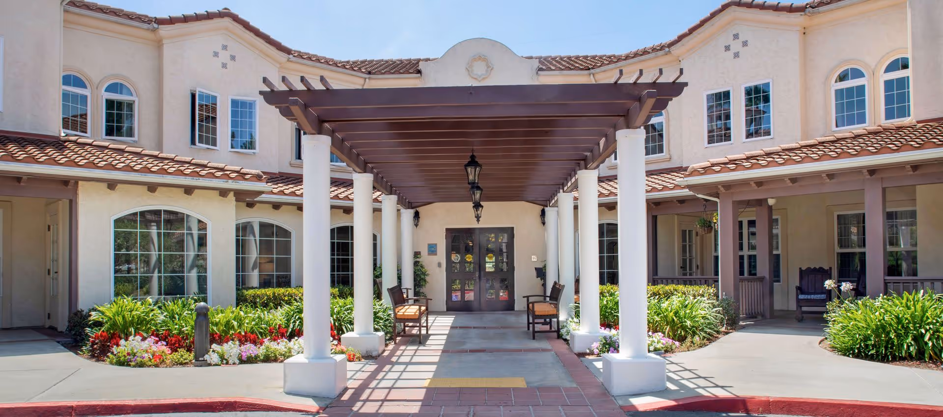 Entrance of a Mediterranean-style senior living building with a pergola supported by white columns, benches, and landscaped flowerbeds.