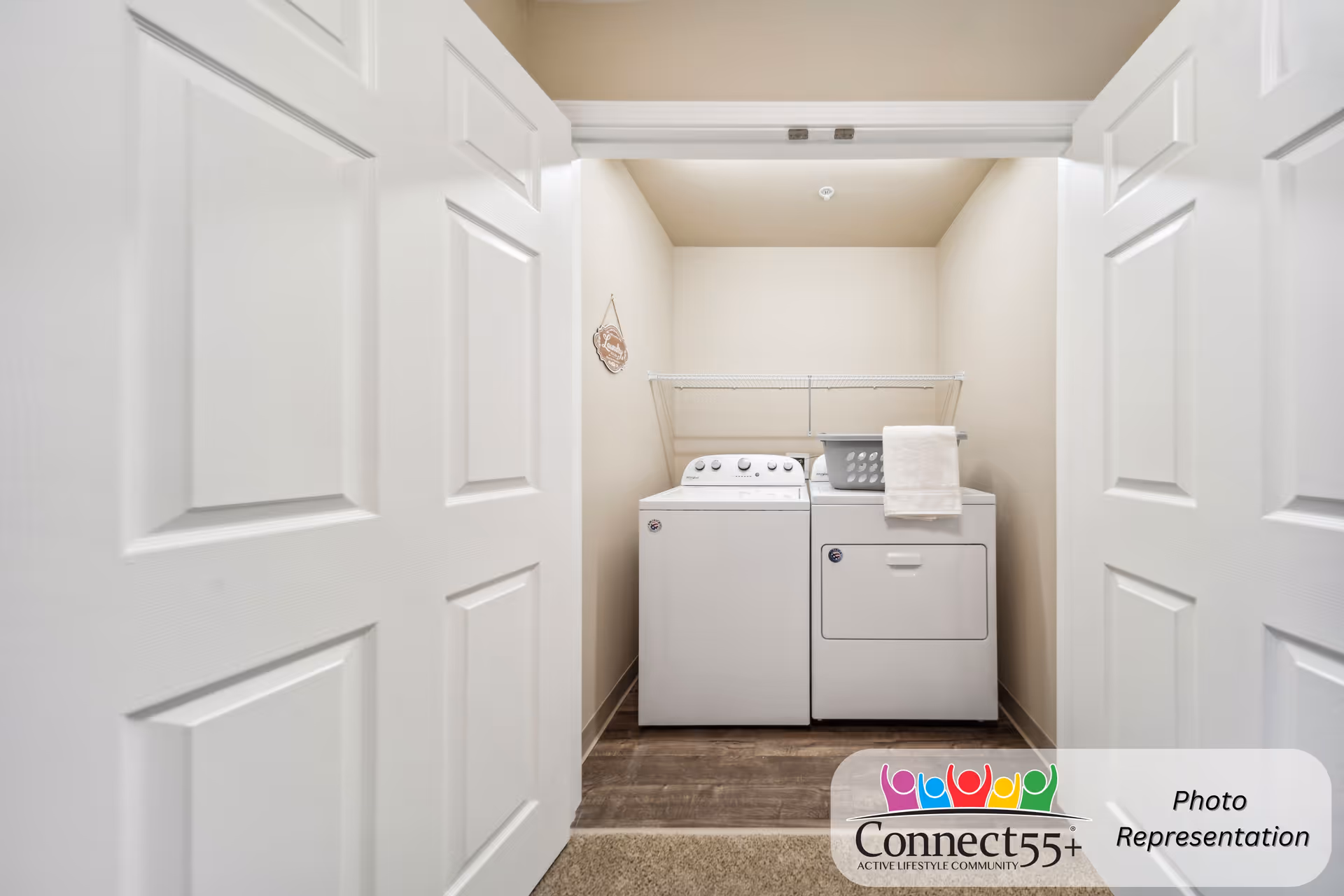 A small laundry room with a white washing machine and dryer side by side, a laundry basket on top of the dryer, and a towel hanging over the dryer. The room has beige walls, a wire shelf above the machines, and white double doors partially open.