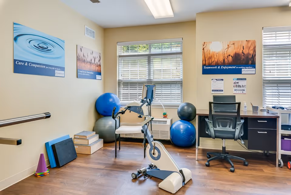 Therapy/fitness room with a stationary exercise bike, stability balls, a desk and chair beneath windows and motivational posters on the walls.