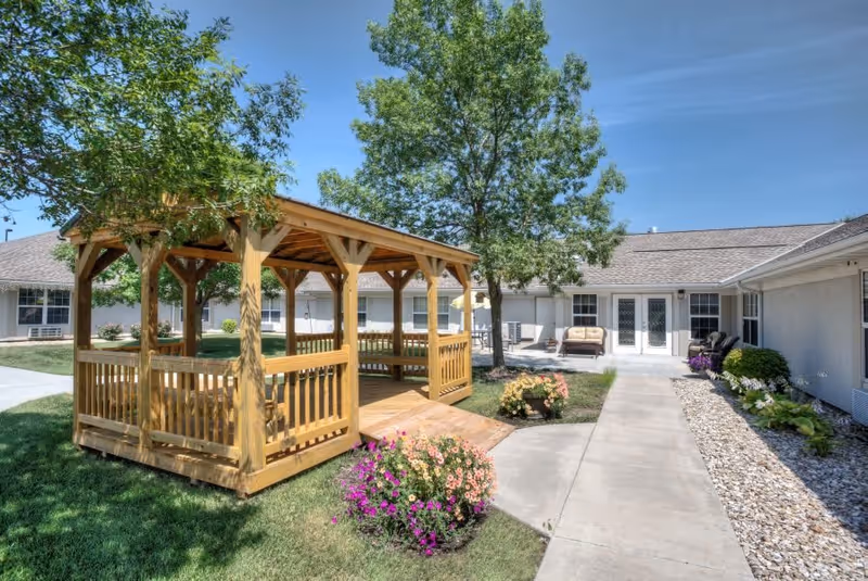 Outdoor courtyard area at Vintage Park at Hiawatha featuring a wooden gazebo surrounded by green grass, colorful flowers, trees, and a concrete walkway leading to a building with multiple doors and windows under a clear blue sky.