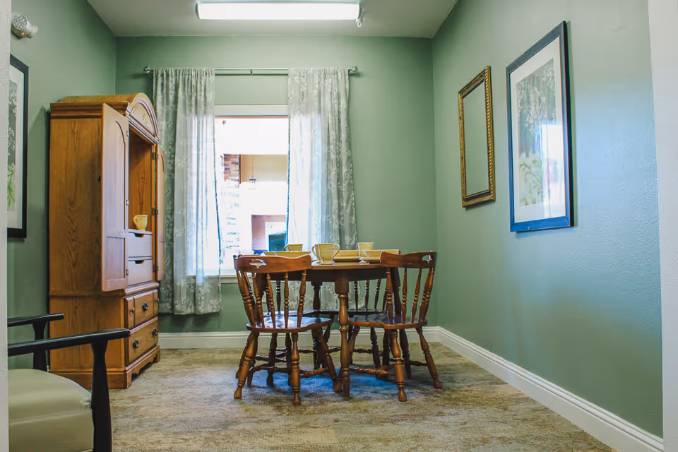 Small dining area with a round wooden table and four chairs in a green-walled room with a wooden cabinet, window with curtains, and framed artwork.