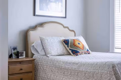 A neatly made bed with a beige upholstered headboard, white and patterned pillows, and a light-colored textured bedspread in a softly lit bedroom. Next to the bed is a wooden nightstand with two framed photos and a small decorative item. A window with white blinds is partially visible on the right side.
