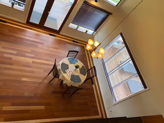 View from above of a small dining area with a round table covered with a white tablecloth and four chairs around it. The table has placemats, a small flower arrangement, and a folded paper. The room has wooden flooring with a decorative border, large windows with blinds, and a hanging light fixture with multiple bulbs.