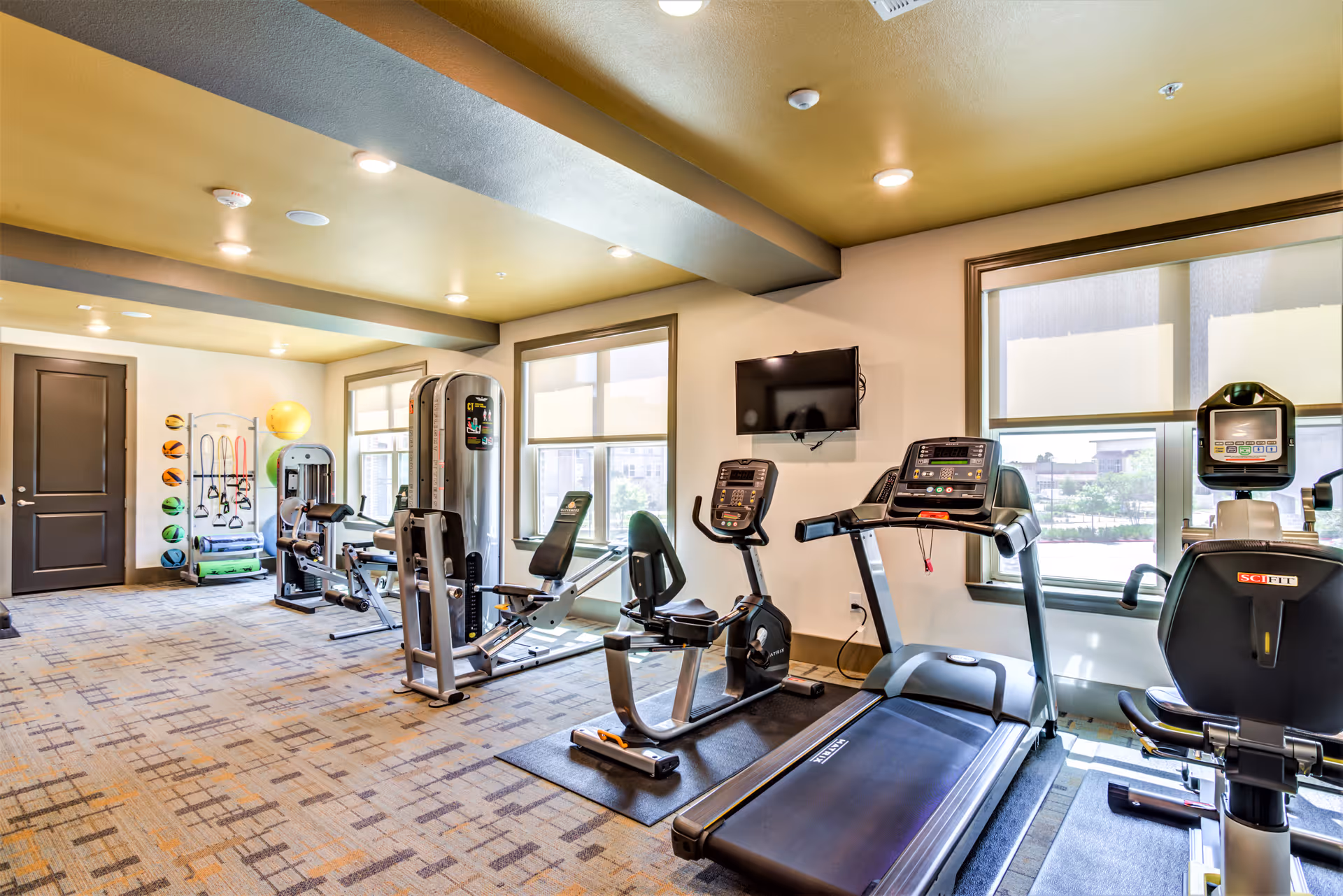 A well-lit fitness room with exercise equipment including treadmills, stationary bikes, weight machines, and various fitness accessories like exercise balls and resistance bands. The room has large windows with blinds, a wall-mounted TV, and patterned carpet flooring.