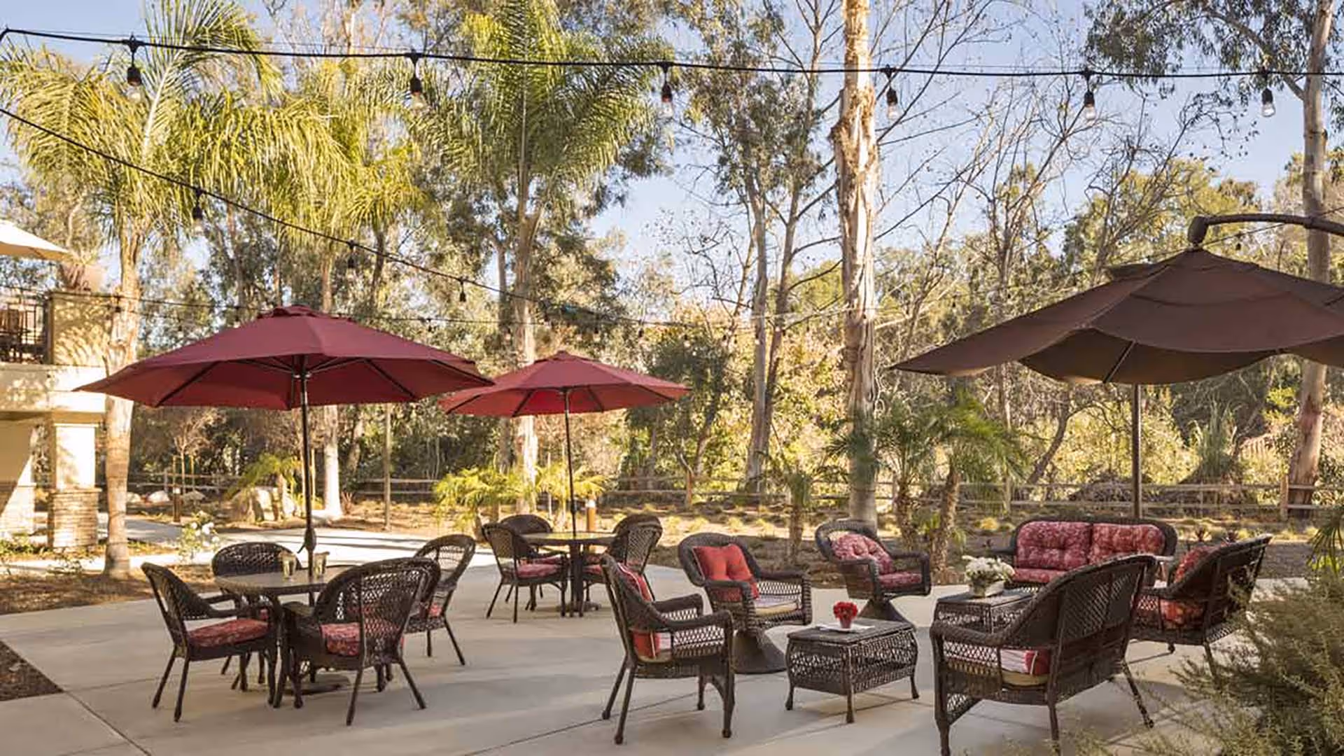 Outdoor patio area with multiple sets of wicker chairs and tables, some with red umbrellas providing shade. The seating includes cushioned chairs and sofas arranged around small tables. The area is surrounded by trees and greenery under a clear sky.