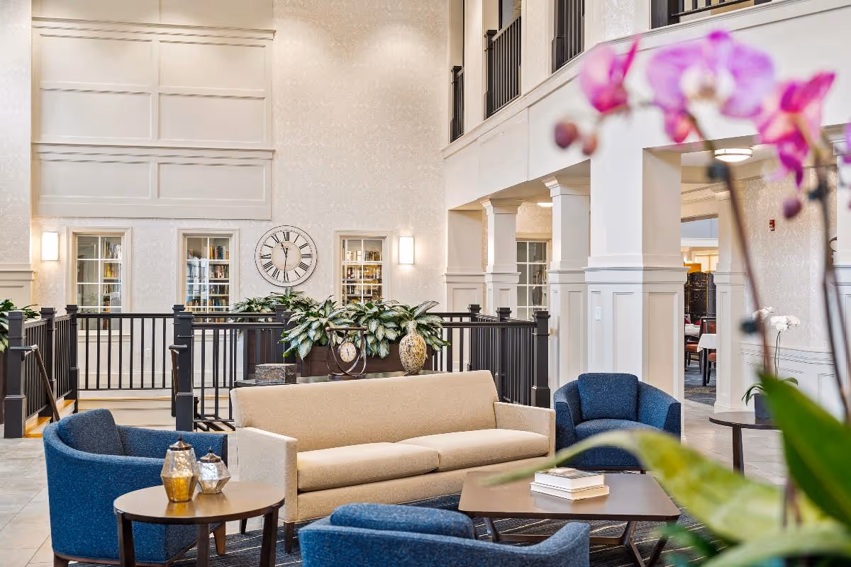 Bright, elegant common lounge with a beige sofa, blue armchairs, coffee tables, plants, and a large wall clock.