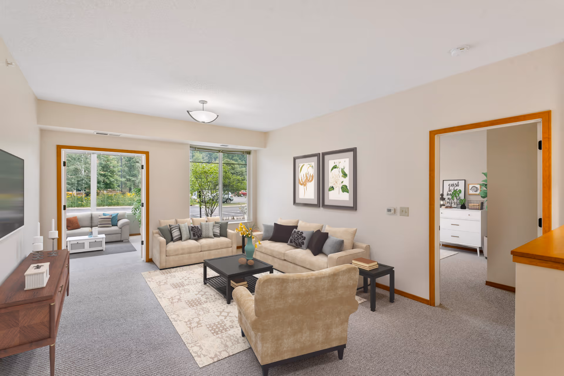 A bright living room with beige sofas, a matching armchair, a black coffee table with a vase of yellow flowers, and two framed botanical prints on the wall. There is a large window letting in natural light and a door leading to a sunroom with additional seating. The room has light-colored walls and carpeted floors.