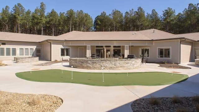 Outdoor courtyard area of a single-story assisted living facility with a putting green in the center, surrounded by a concrete walkway and a stone circular seating or fire pit area. The building has beige siding and a brown roof, with trees in the background.