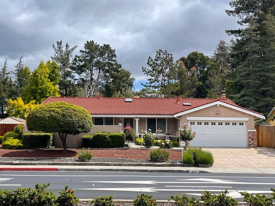 Single-story residential building with a red tiled roof, beige exterior walls, and a white garage door. The front yard is landscaped with trimmed bushes, a uniquely shaped tree, and various plants. The sky is cloudy and there are tall trees in the background.