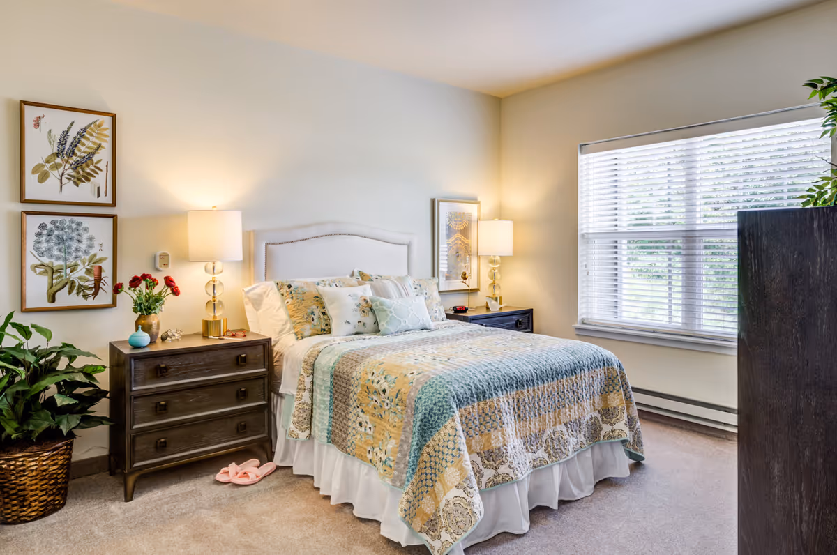 A cozy bedroom with a neatly made bed featuring a patterned quilt and multiple pillows. There are two dark wooden nightstands on either side of the bed, each with a lamp. The room has light-colored walls, a large window with white blinds, framed botanical artwork on the wall, a potted plant in a woven basket, and a pair of pink slippers on the carpeted floor.