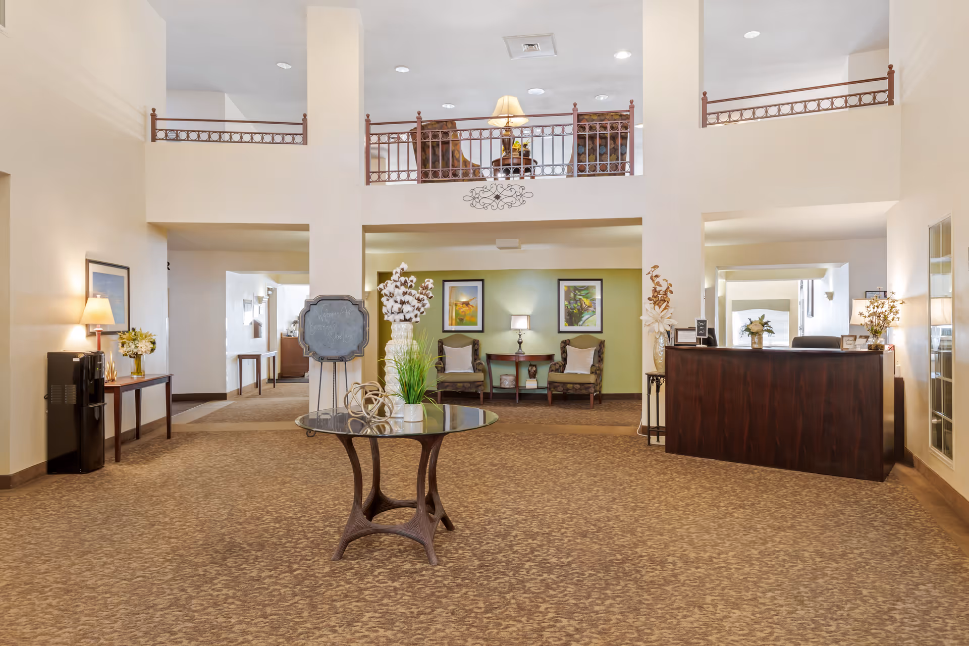 Spacious lobby area with beige walls and carpet, featuring a round glass table with decorative plants and sculptures in the center. To the right is a dark wooden reception desk with lamps and flower arrangements. Behind the table, there are two armchairs with a small table and lamp between them against a green accent wall with framed artwork. The upper level has a balcony with two patterned chairs and a lamp on a small table.