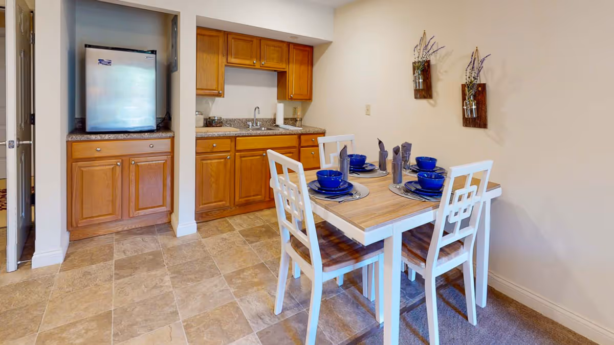 A small dining area with a wooden table set for four with blue bowls and napkins. The dining area is adjacent to a kitchenette with wooden cabinets, a small refrigerator, a sink, and a granite countertop. Two decorative wall hangings with flowers are mounted on the wall above the dining table.