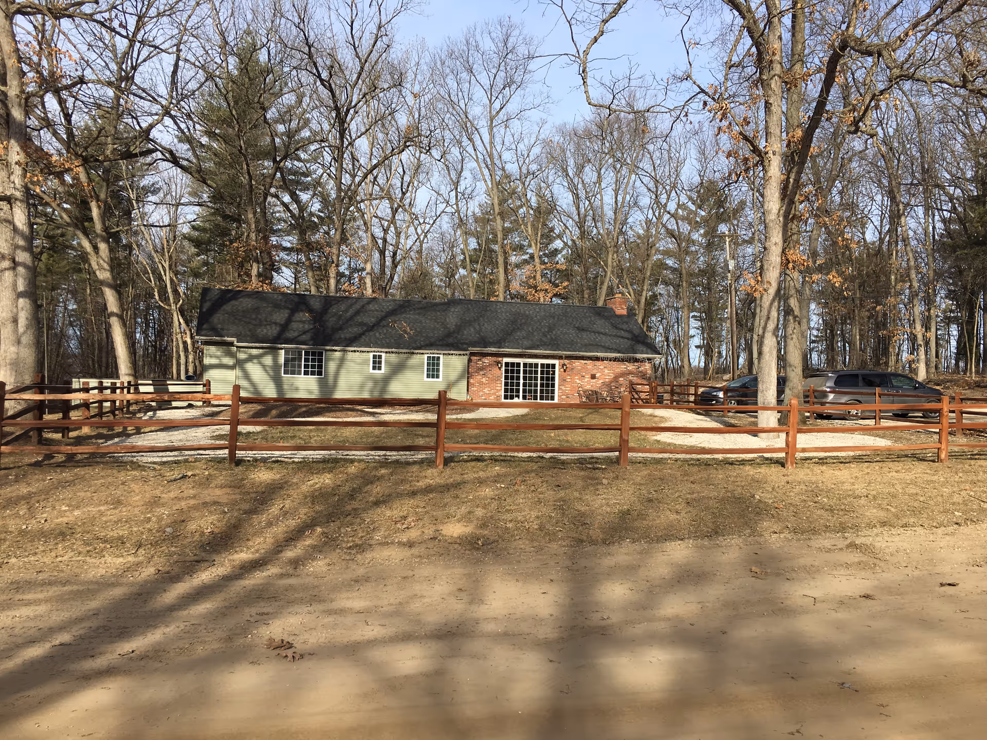 A single-story house with green siding and a brick chimney, surrounded by a wooden fence. There are two vehicles parked on the right side of the house. The house is set against a backdrop of leafless trees under a clear blue sky.