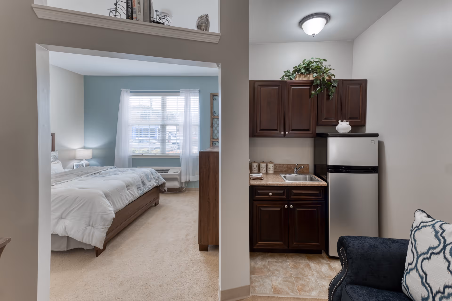 View of a senior living facility room showing a bedroom with a bed, nightstand, lamp, and window with sheer curtains on the left, and a small kitchenette area with dark wood cabinets, a sink, countertop, and a mini refrigerator on the right. A blue upholstered chair with a patterned pillow is partially visible in the foreground.