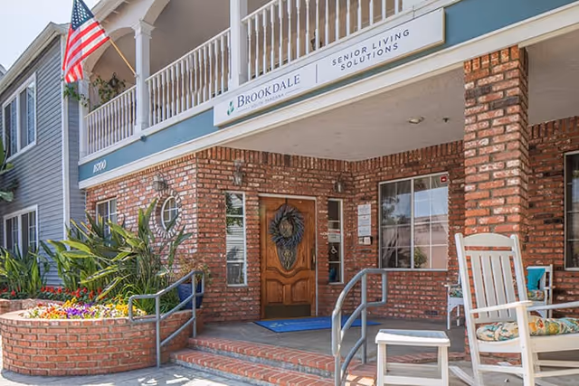 Entrance of Brookdale Gardens of Tarzana senior living facility featuring a brick facade, a wooden door with a decorative wreath, an American flag, potted plants, and a white rocking chair on the porch.