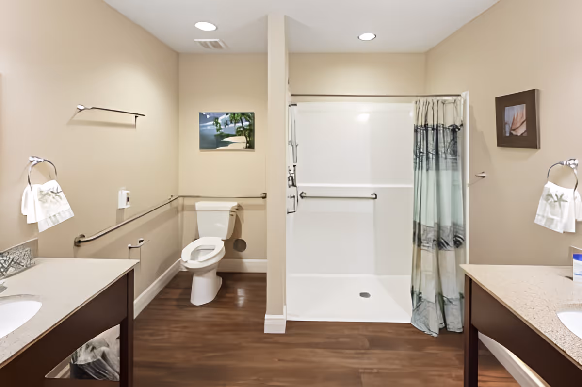 A clean and accessible bathroom featuring a toilet with grab bars, a walk-in shower with a curtain, two sinks with countertops on either side, towel holders with white towels, and framed pictures on the beige walls. The floor is wooden, and the room is well-lit with recessed ceiling lights.
