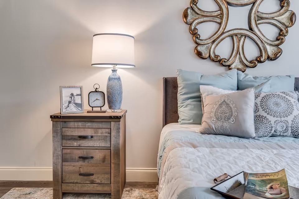 A bedside table with a lamp, clock, and framed photo next to a neatly made bed with decorative pillows and wall art.