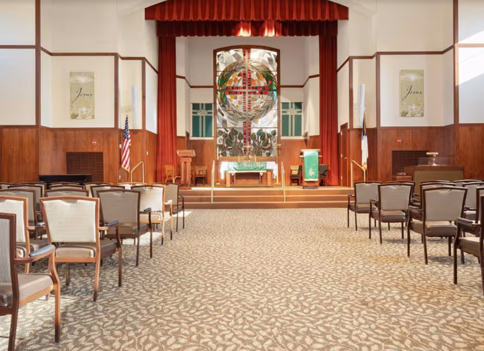 Interior view of a chapel with rows of chairs facing a raised altar area. The altar features a large stained glass window with a cross design, flanked by red curtains. There are two podiums and religious banners on either side of the altar. The walls are wood-paneled halfway up with light-colored paint above, and the carpet has a leaf pattern.