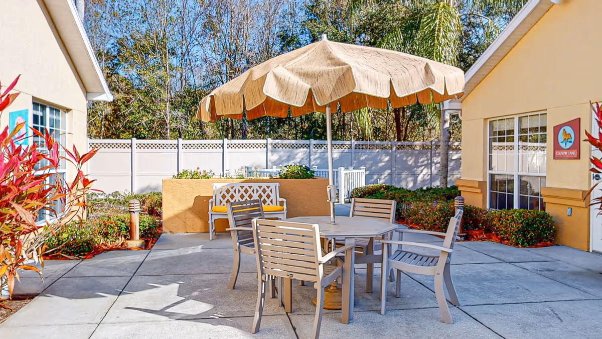 Outdoor patio area with a round table and four chairs under a large beige umbrella. There is a white bench with a yellow cushion against a low wall, surrounded by bushes and plants. The area is enclosed by a white fence, with two beige buildings on either side and trees in the background.