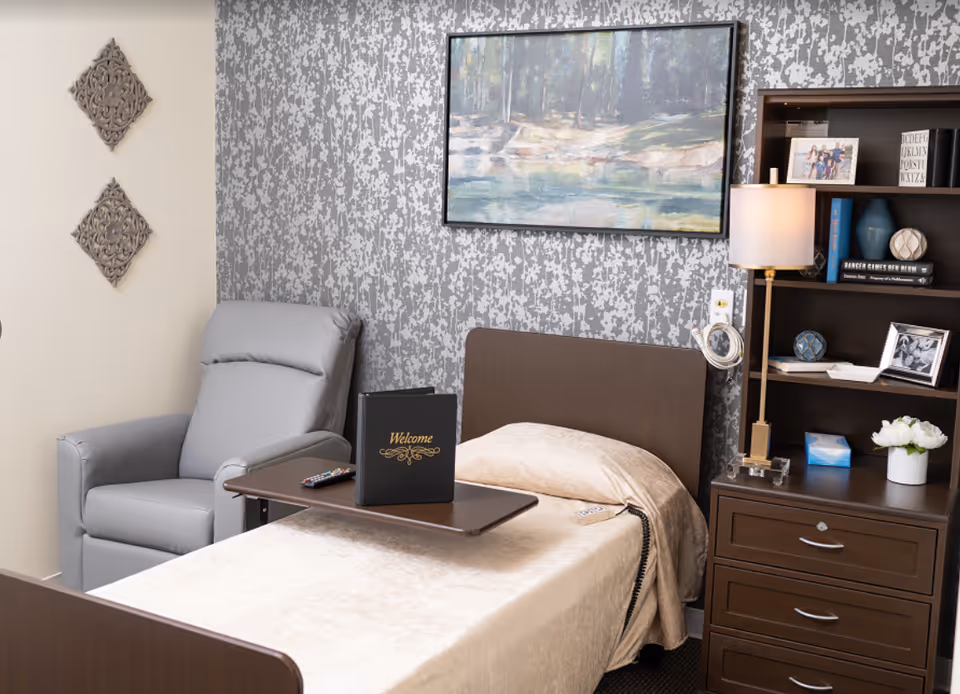 Furnished senior living bedroom with a single bed, overbed table with a welcome binder, gray recliner, bedside dresser, lamp and shelving.