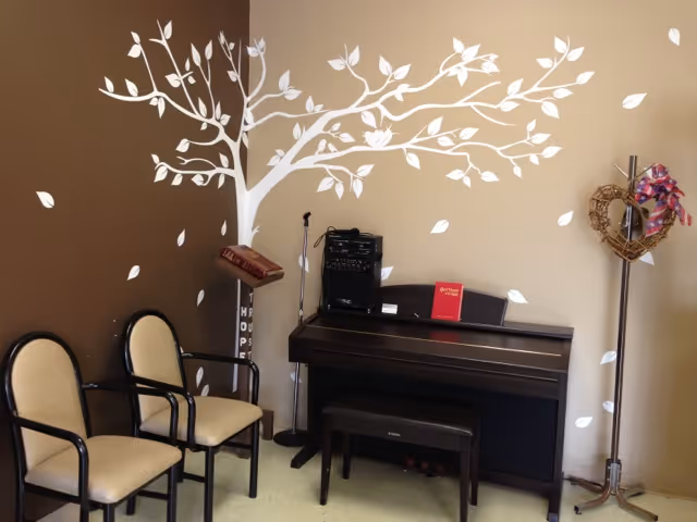 A small room with two beige chairs with black frames, a black upright piano with a matching bench, a microphone stand, and a music stand. The walls are painted brown and beige with a white tree decal spreading across the corner. A heart-shaped wreath with a pink ribbon hangs on a metal coat rack.