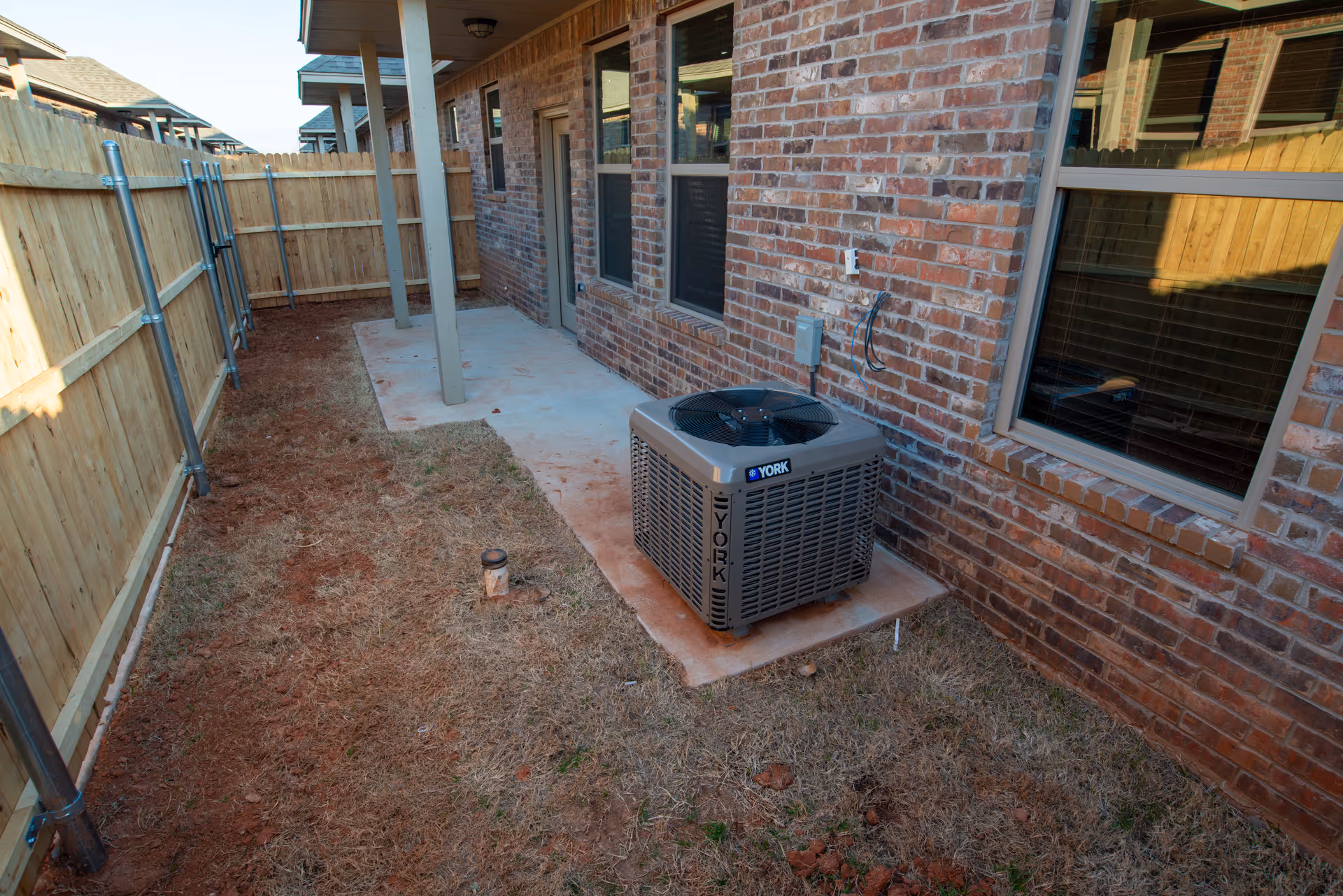 Narrow fenced side yard with a concrete patio, central air unit by a brick house and several windows.