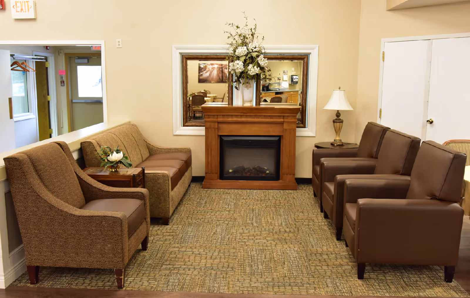 A cozy sitting area in an assisted living facility featuring a wooden electric fireplace with a floral arrangement on top, a large mirror behind it, two brown upholstered armchairs on the right, and a patterned loveseat and armchair on the left with a small side table holding a flower arrangement. A table lamp is on a small table next to the armchairs, and a hallway with a door and coat hangers is visible in the background.