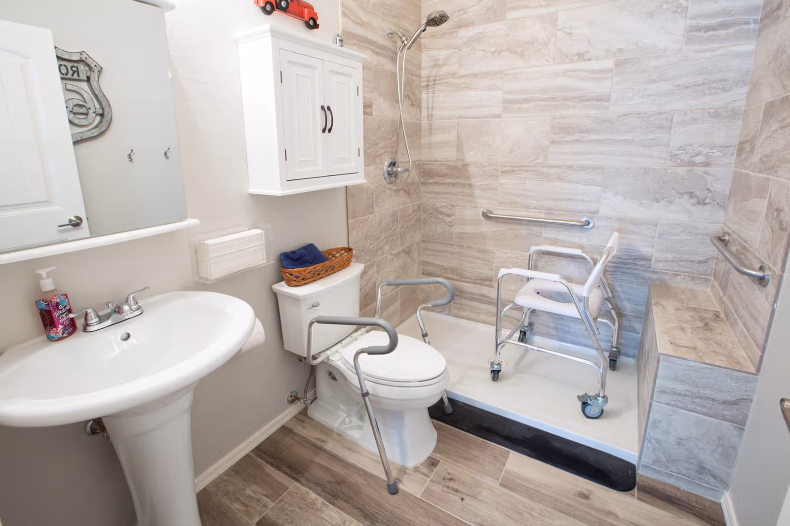A bathroom designed for accessibility with a pedestal sink, a toilet equipped with safety rails, and a walk-in shower area featuring a shower chair with wheels and multiple grab bars on the tiled walls. The floor has wood-like tiles and there is a white cabinet mounted on the wall above the toilet.