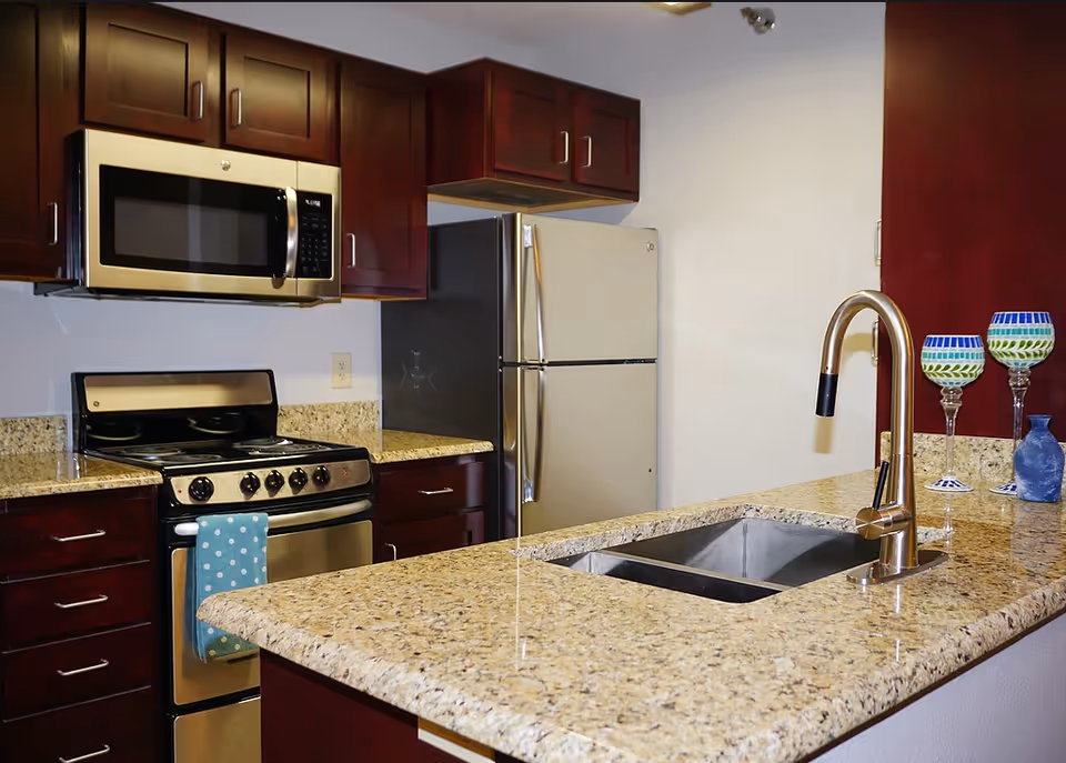 Modern kitchen with granite countertops, stainless steel appliances including a microwave, stove, and refrigerator. There is a double sink with a curved faucet on the countertop, and two decorative glasses and a small blue vase are placed on the counter.