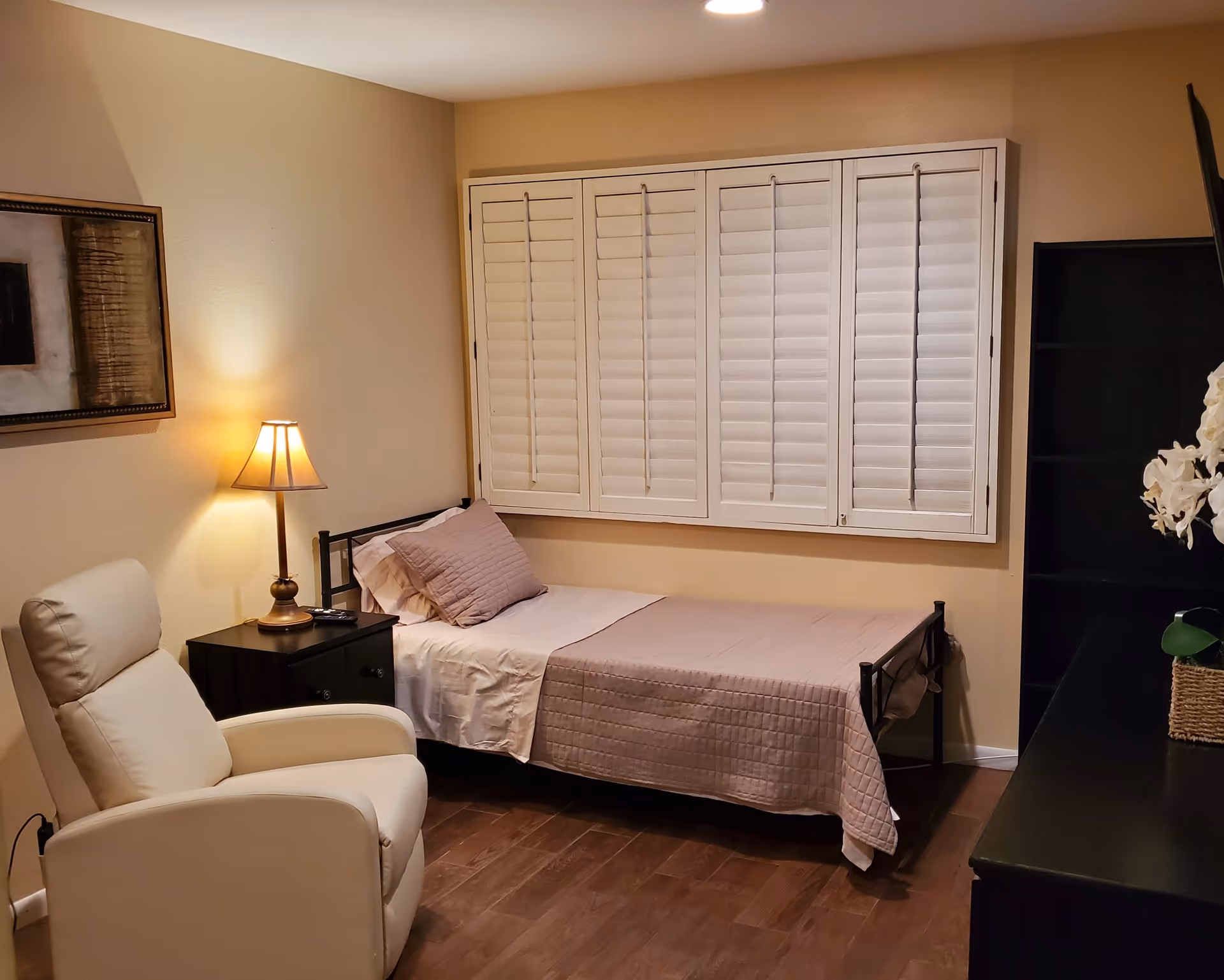 A small bedroom with a single bed covered with a light purple quilt and pillow. Next to the bed is a black nightstand with a lamp and a remote control. A white recliner chair is positioned near the nightstand. The room has wooden flooring, beige walls, a framed abstract painting, and white plantation shutters covering the window. There is also a black shelving unit and a TV on a black stand with a white flower arrangement.
