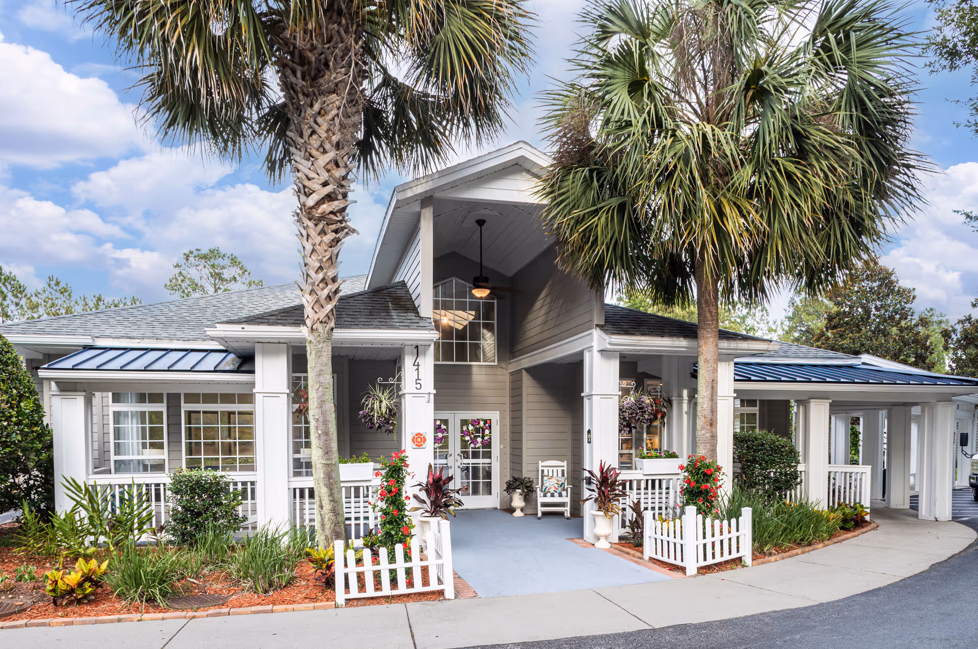 Front entrance of a one-story building with a covered porch, two palm trees, white railings, picket fences and landscaped beds.
