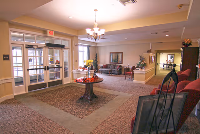 Interior view of a senior living facility lobby area with a round table decorated with flowers and a bowl of red ornaments in the center. There are glass double doors leading outside, a chandelier hanging from the ceiling, and seating areas with chairs and a sofa along the walls. The space is carpeted and well-lit with natural light coming through the windows.