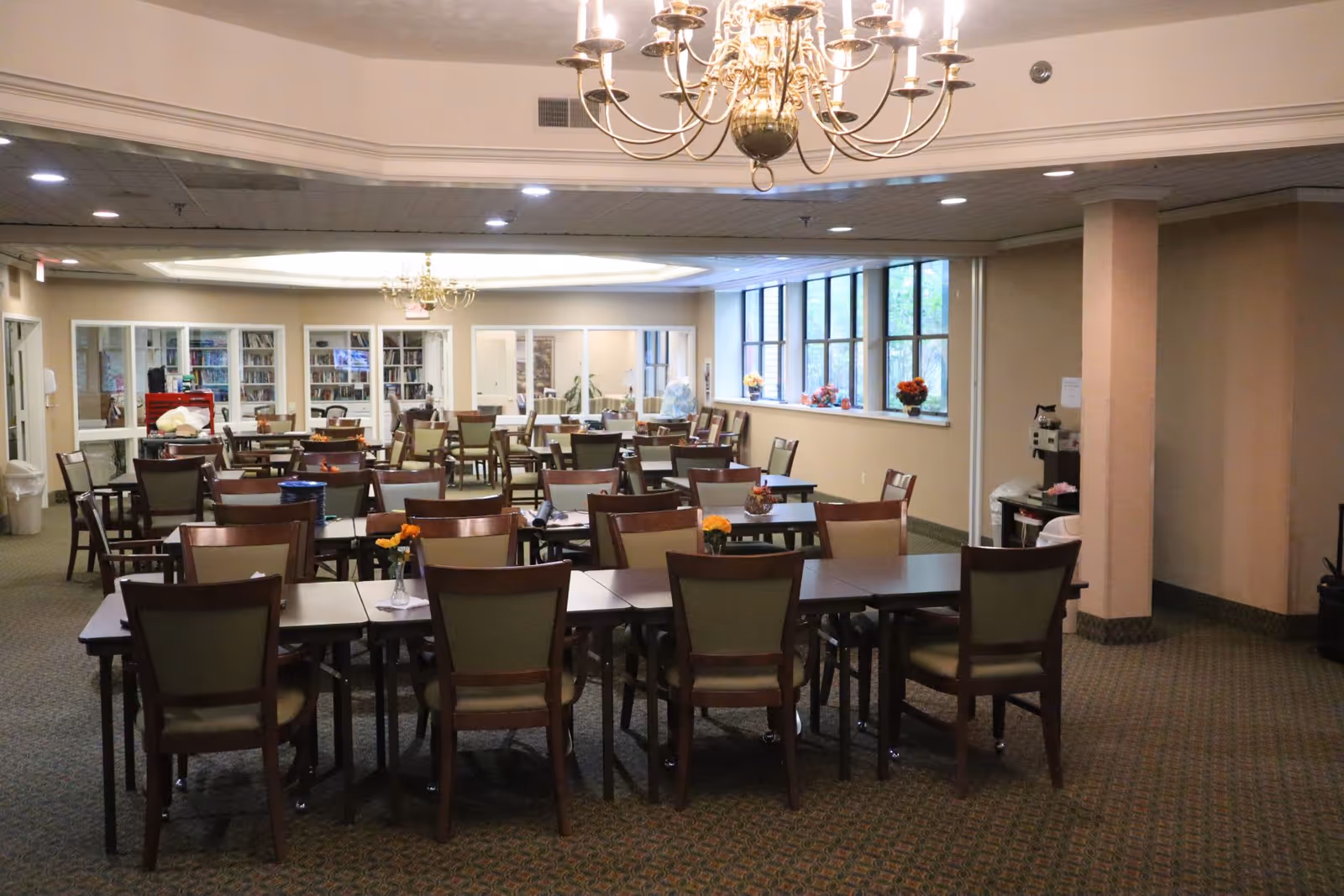 A spacious dining room with multiple wooden tables and chairs arranged neatly. The room features large windows on one side letting in natural light, chandeliers hanging from the ceiling, and a carpeted floor. There are flowers in small vases on some tables and a bookshelf visible in the background.