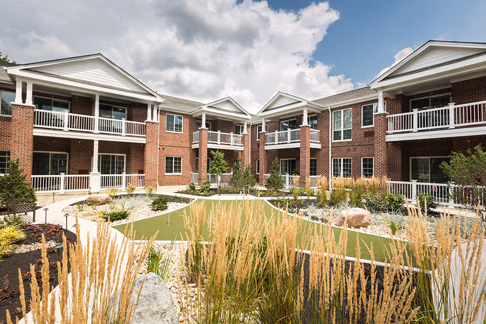 Outdoor courtyard area of a senior living facility with a putting green, landscaped garden beds, benches, and a two-story brick building with white balconies surrounding the courtyard under a partly cloudy sky.