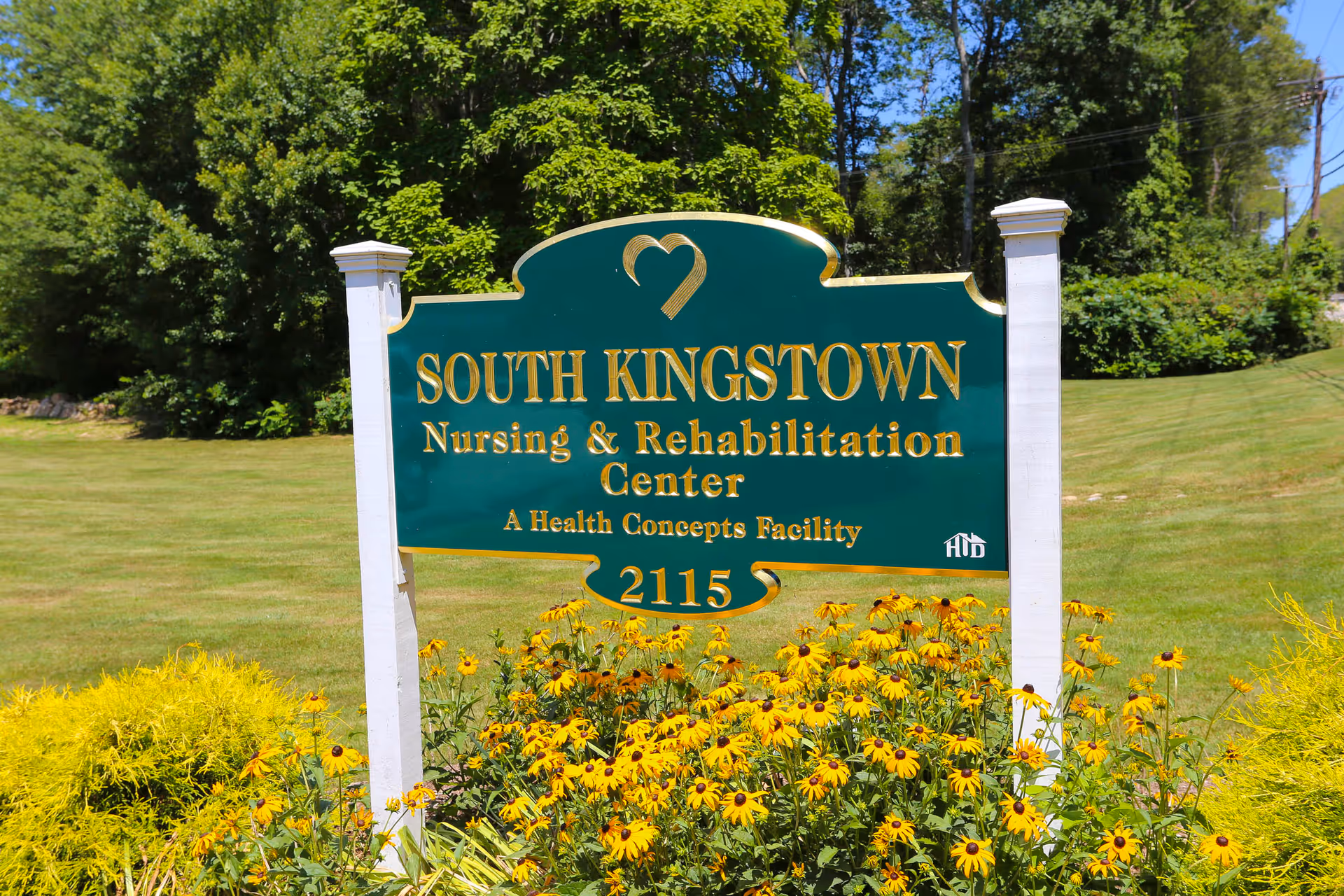 Green and gold sign for South Kingstown Nursing & Rehabilitation Center surrounded by yellow flowers and a grassy lawn.