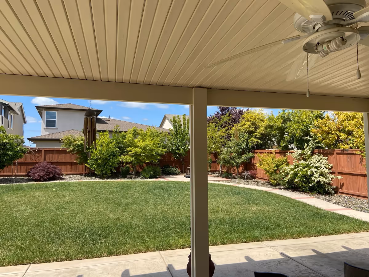 View from a covered patio with a ceiling fan and wind chime looking out onto a green lawn, shrubs and a wooden fence under a blue sky.