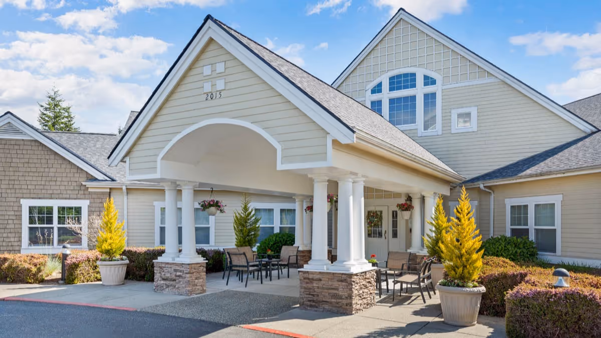 Covered porte-cochère entrance of a beige senior living building with columns, outdoor seating, and potted plants.