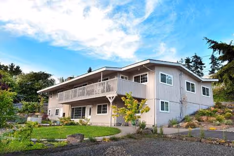 Two-story light-gray senior living facility with balcony, landscaped yard, and curved driveway.