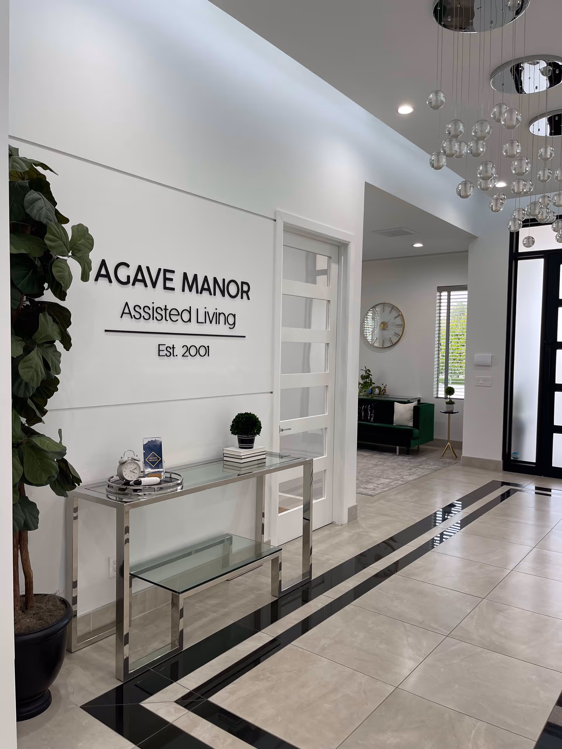 Interior view of the entrance area of Agave Manor Assisted Living facility. The wall displays the facility name and establishment year. There is a modern glass and metal console table with decorative items including a small potted plant, books, and a clock. A large potted plant is on the left side. The floor has a polished tile design with black accents. In the background, a seating area with a green couch, a round wall clock, and a window with blinds is visible. A modern chandelier with hanging glass orbs is seen on the ceiling.
