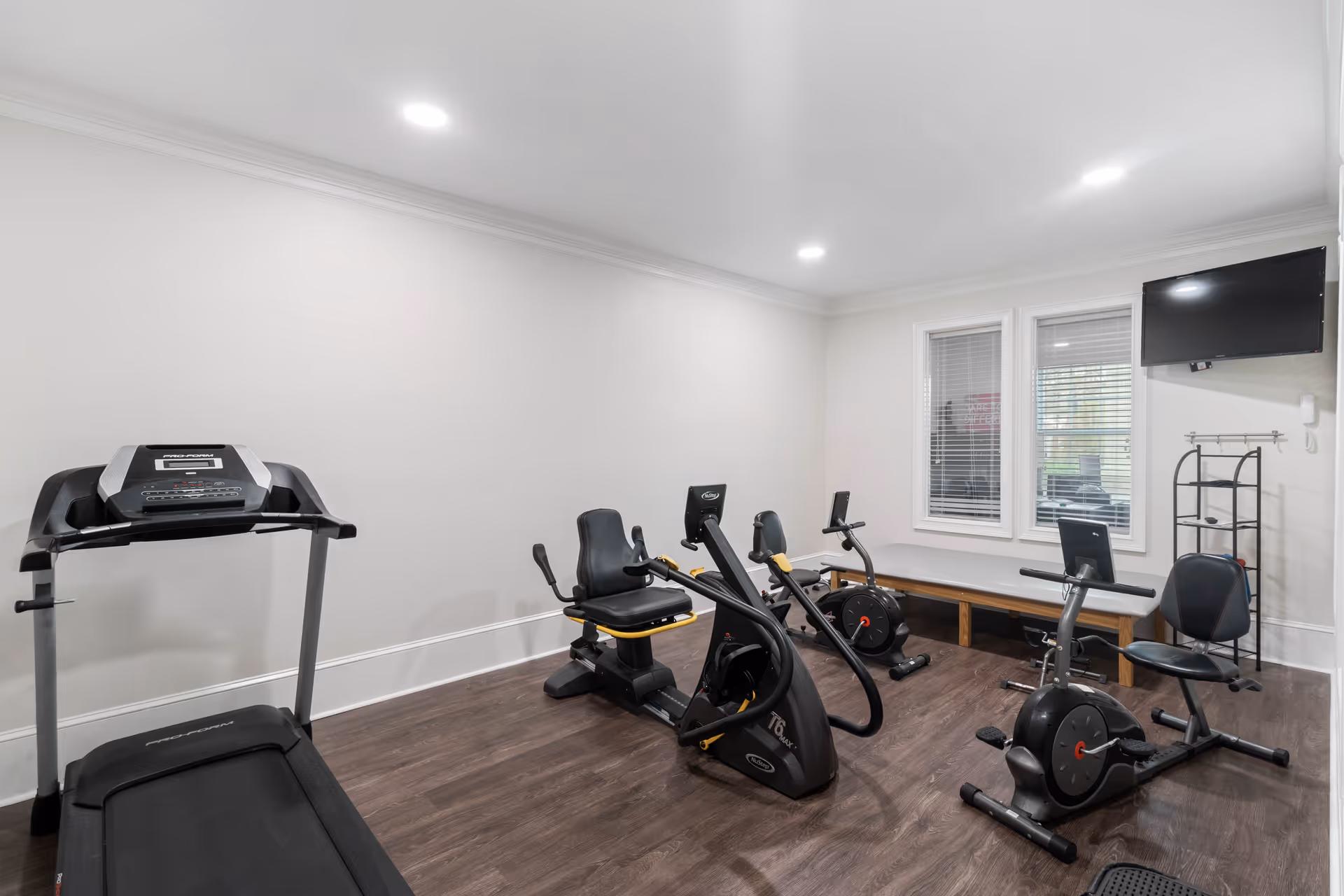 A small fitness room with exercise equipment including a treadmill, two recumbent stationary bikes, and a rowing machine. The room has white walls, wood flooring, two windows with blinds, a wall-mounted TV, and a bench along the wall.