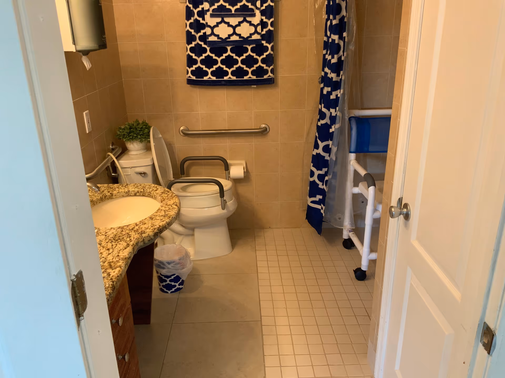 A bathroom with beige tiled walls and floor, featuring a toilet with safety rails, a granite countertop sink, a blue and white patterned towel hanging on the wall, a matching shower curtain, and a shower chair inside the shower area.