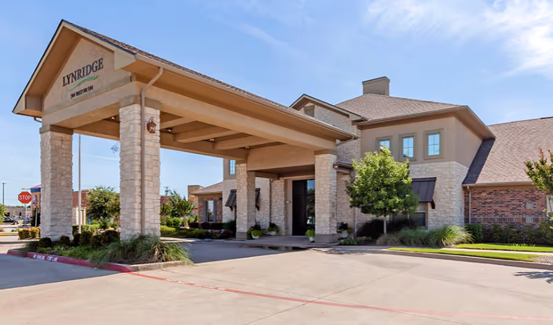 Exterior view of Lynridge of Murphy Assisted Living & Memory Care building with a covered entrance supported by stone pillars, surrounded by landscaping and a clear blue sky.