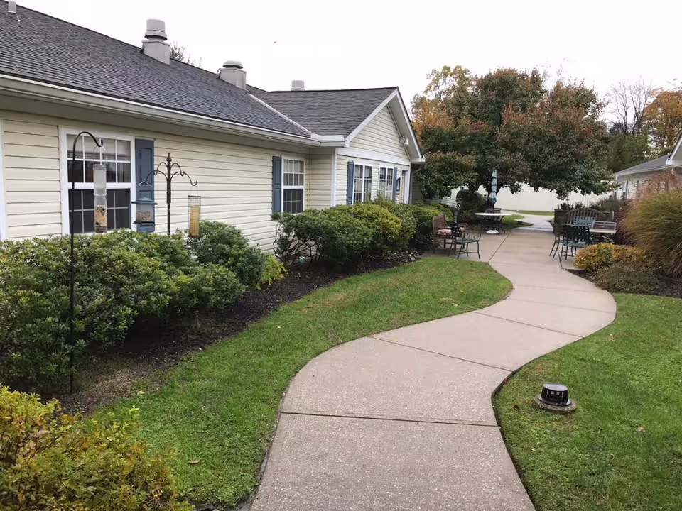 A winding concrete pathway runs through a well-maintained garden area with green grass and bushes. On the left side, there is a beige building with blue shutters and multiple windows. Bird feeders hang on metal poles near the building. On the right side, there are several outdoor chairs and tables under a large tree with autumn-colored leaves.